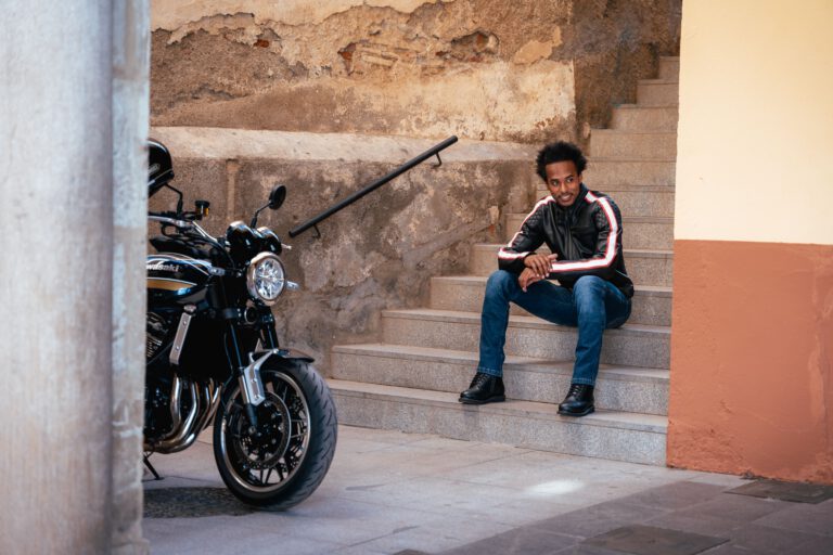 Ein Mann mit Jeans und Lederjacke sitzt auf einer Treppe, vor ihm steht ein Motorrad - Chris Gollhofer Fashion Fotografie